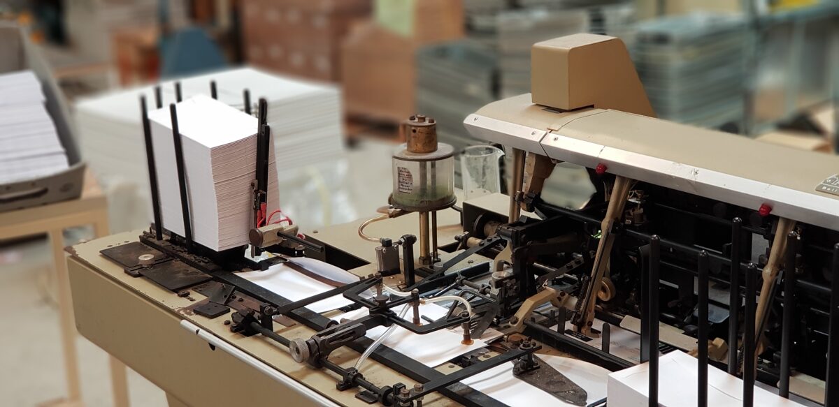 A mail inserting machine on the floor at Direct Print and Mail Solutions, a stack of printed sheets loaded in the feed tower and the folding and inserting mechanism running alongside it.
